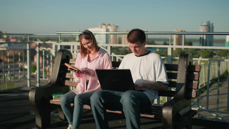 young adults seated outdoors on a wooden bench, male typing on laptop while focused, female reading from book with serene background featuring modern buildings, and greenery