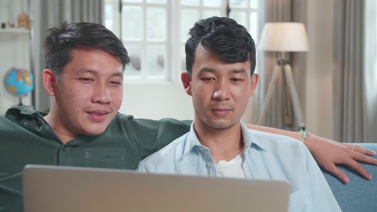 Asian Gentle Gay Couple Using Laptop Computer, While Sitting On A Couch In Cozy Stylish Apartment. Adult Boyfriends Online Shopping On Internet, Watching Funny Videos On Streaming Service