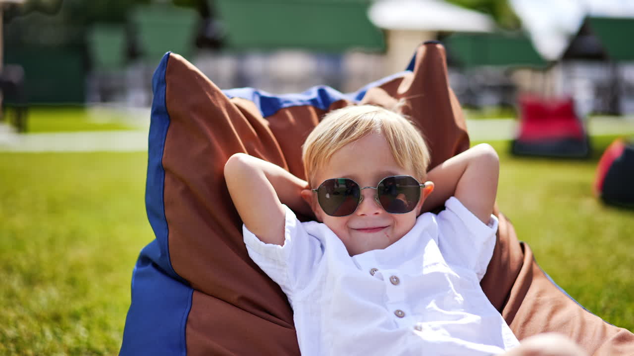 Funny blond little boy wearing big sunglasses spends time outdoors. Cute kid folded his arms behind the head lying in a comfortable chair.