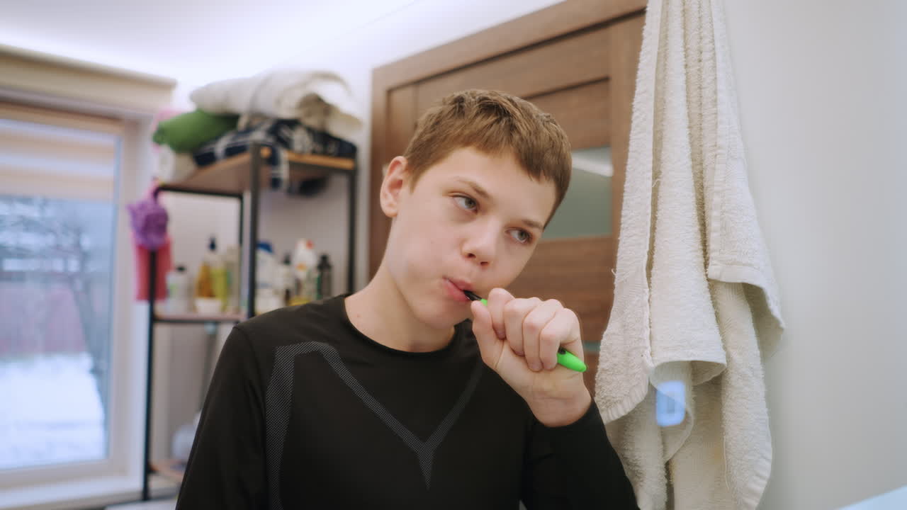 Fokussierter weißer Enkel putzt Zähne am Waschbecken, Nahaufnahme Spiegelwinkel zeigen konzentrierten Gesichtsausdruck des Teenagers, detaillierter Minzschaum und Zahnpasta, Handtuch und Fenster im Hintergrund, intime Pflegeszene in der Familie