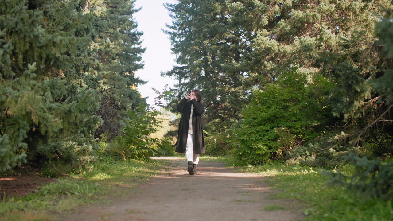 긴 겨울 코트를 입고 수목원을 거닐며, 휴대폰을 들고 키 큰 소나무 아래에서 사진을 찍는 쾌활한 여성, 자연 경관과 초목을 담으며 평온하게 걷고 있어요.
