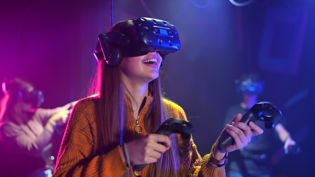 Una bambina felice e interessata gioca su un tapis roulant VR con casco e controller. Altre persone giocano nelle vicinanze. Slow motion