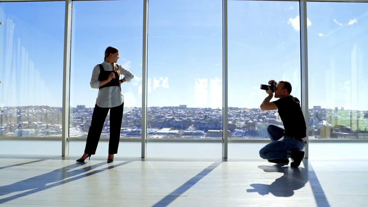 Attractive model in business suit posing on camera. Male photographer taking photos of a young woman in glasses in office with large windows in the city.