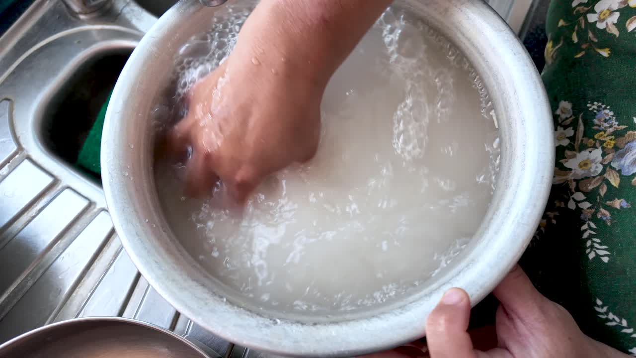 Woman washing rice in metal bowl in kitchen sink, preparing ingredients for cooking