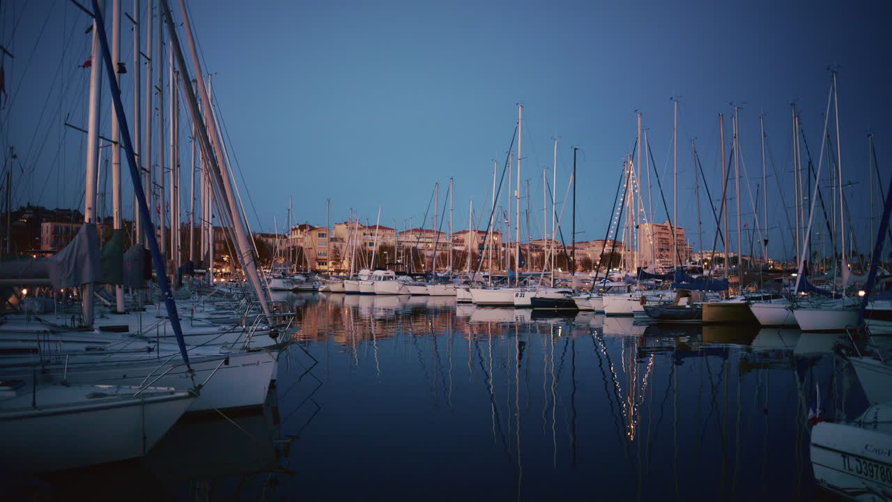 Vidvinkelbild av en hamn i en sydfransk kuststad i skymningen. Båtar ligger stilla på vattnet medan varma byggnadsljus reflekteras på ytan