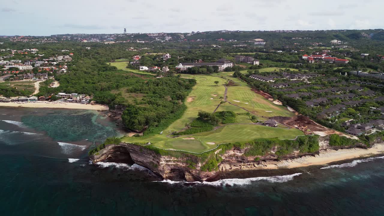 Luxury golf course on a coastal cliff filmed from above showing green fairways sand bunkers resort buildings access roads and ocean waves crashing directly against the rocky shoreline