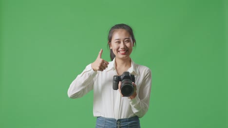 asian photographer with a camera showing thumbs up gesture and smiling while standing on green screen background in the studio