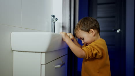 adorable smiling toddler stands holding by the sink in the bathroom. baby boy opens the tap and outs his hand under the water.