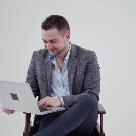 portrait of a handsome businessman with a laptop. smiling young man in grey costume sitting on the chair on the white background. slow motion.
