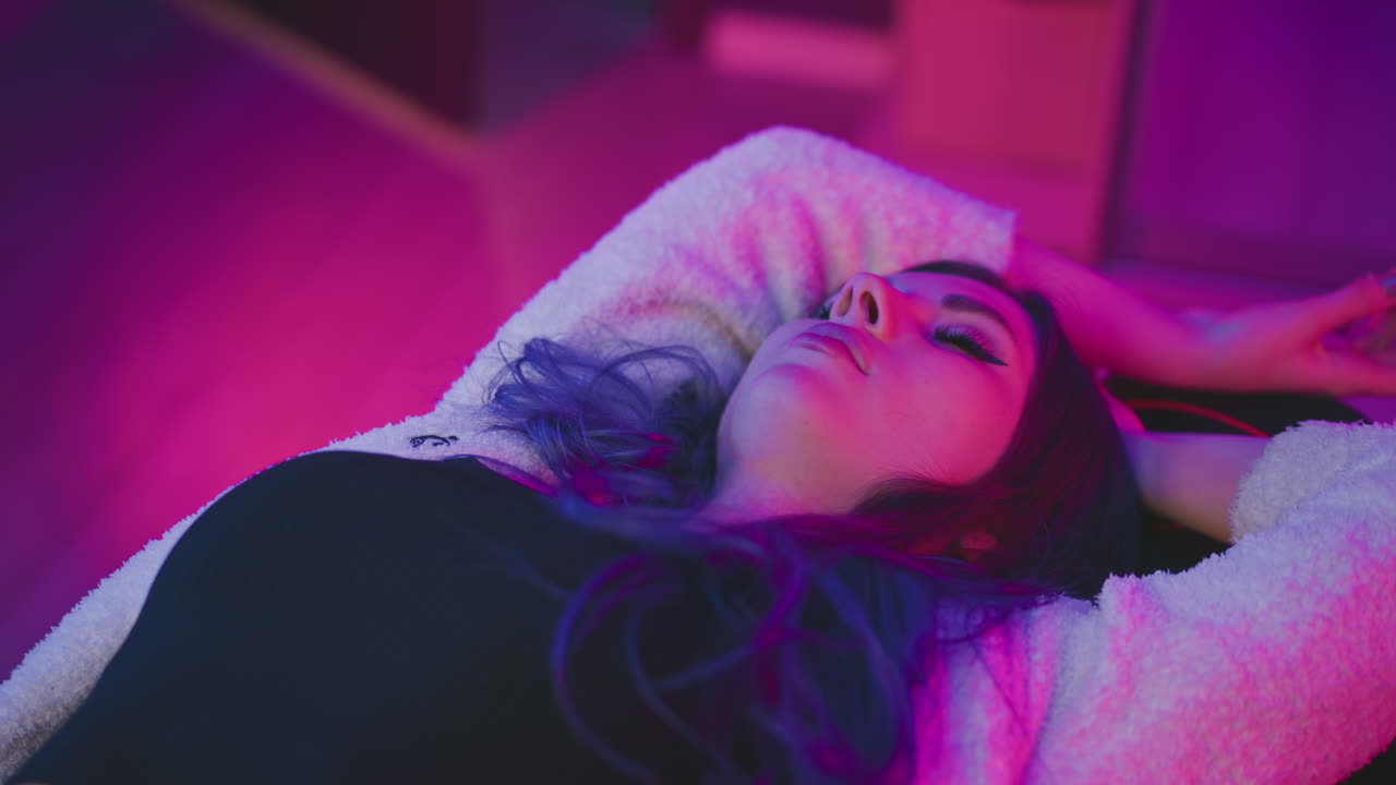 Young woman lying flat asleep with arms over head on soft surface, illuminated by vibrant pink and purple lighting, creating peaceful yet artistic indoor atmosphere with dreamy cinematic mood