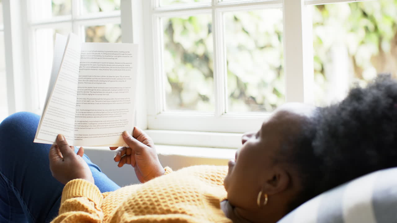 Happy african american senior woman lying, reading book and smiling in sunny room, slow motion
