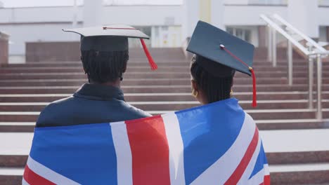 african american man and woman standing side by side with their backs to the camera.they have the british flag on their shoulders. a university building on background