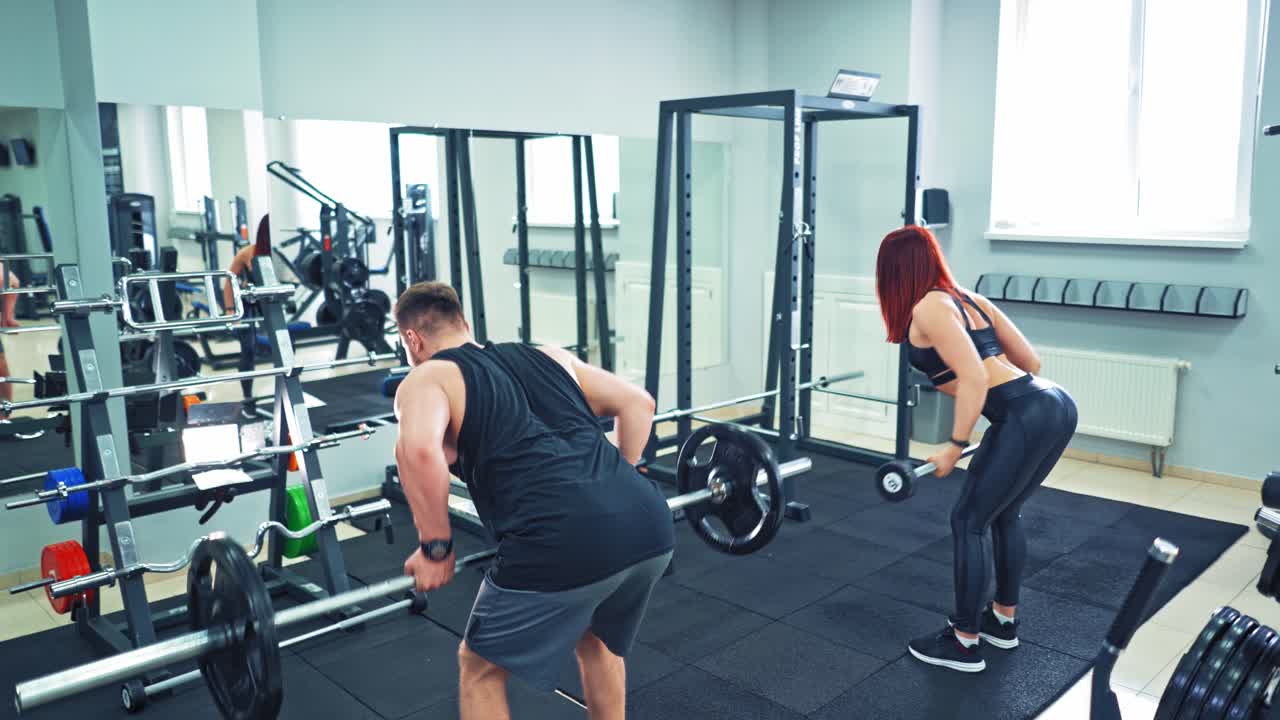 Back view of athletic man and woman lifting barbells in the modern gym. Strong couple is doing physical exercises with heavy equipment standing in front of a big mirror in fitness club.