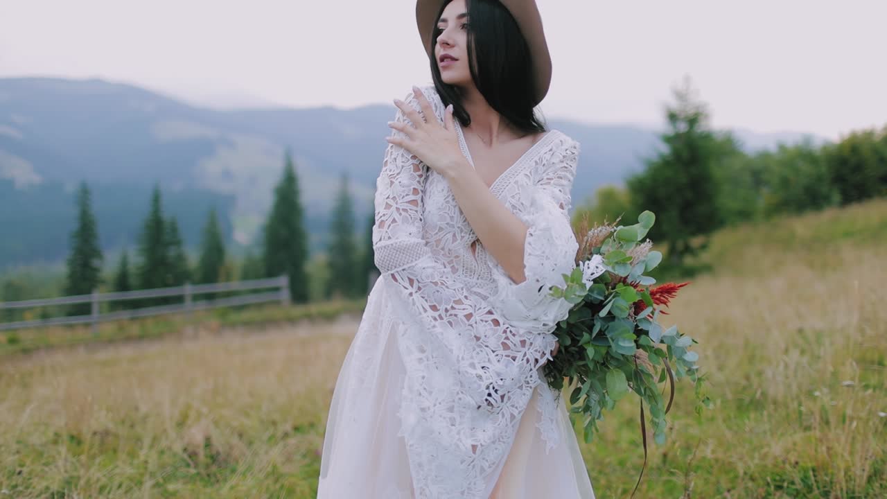 Portrait of a luxury girl in the mountains. Attractive brunette model in beautiful dress and hat embracing herself while standing in nature.