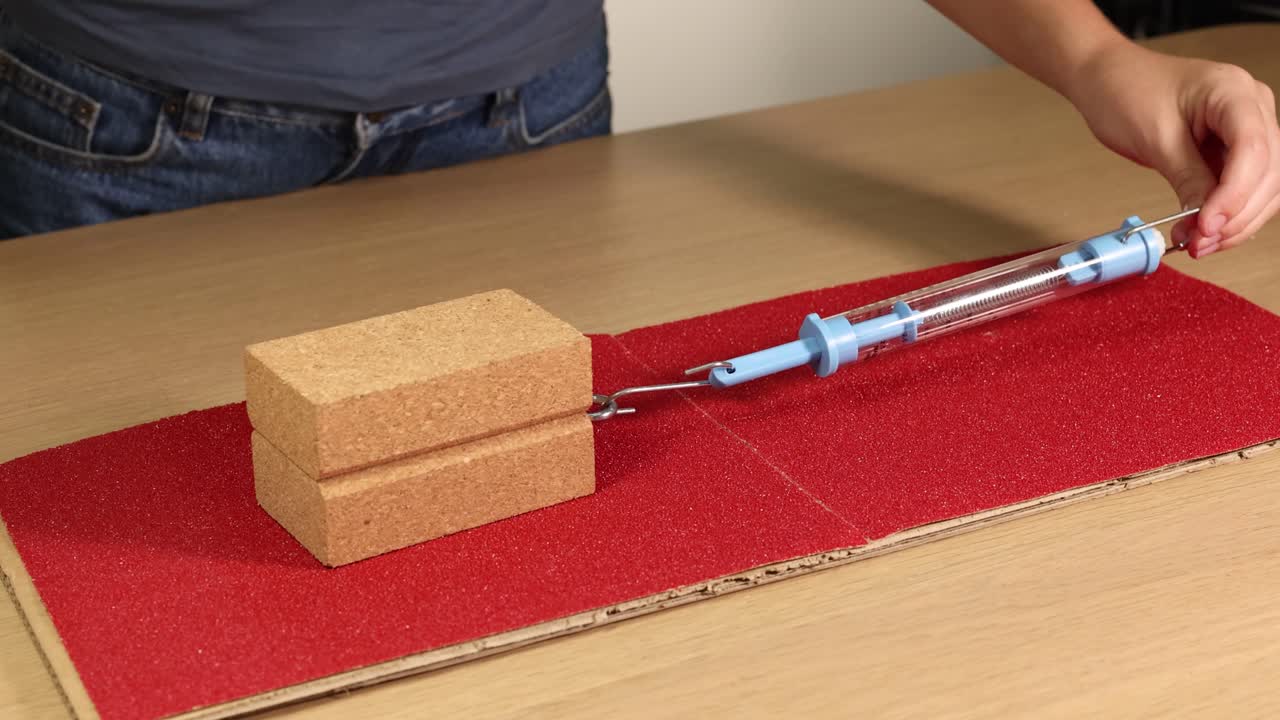 A wooden block is dragged across sandpaper using a spring scale, illustrating friction and force measurement