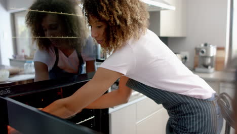 happy biracial mother putting cakes in oven, baking with daughter in kitchen, slow motion