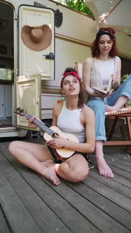 two young women playing ukulele on a wooden deck by a camper van