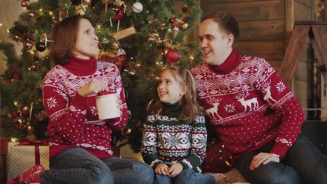 happy mother, father and little daughter eating cookies and kissing each other while sitting in front of christmas tree at home