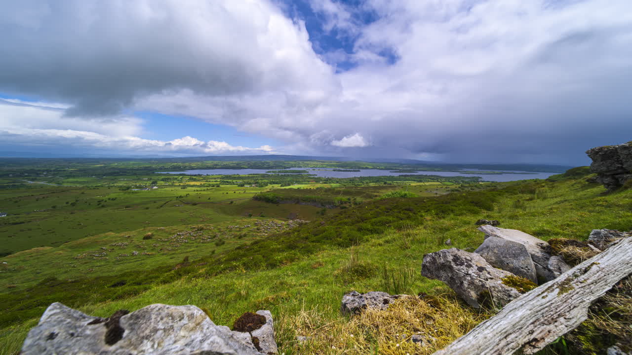 Tidsforløp av en landlig landskapsdal med en passerende regnbyge, med åser og en innsjø i det fjerne under en overskyet, solrik vårdag, sett fra Carrowkeel i Sligo fylke i Irland.