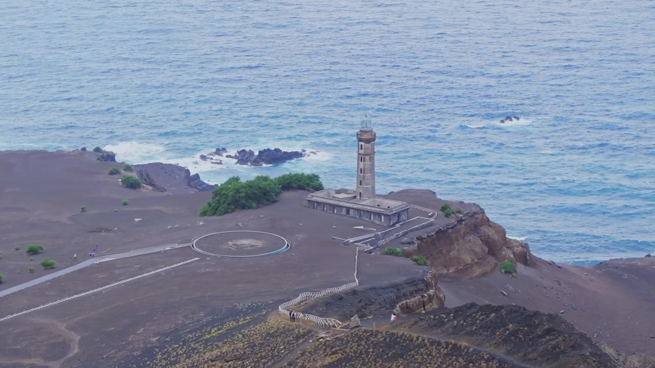 The stone lighthouse of Capelinhos stands alone on a wide grey field of volcanic ash above a blue ocean on Faial Island.