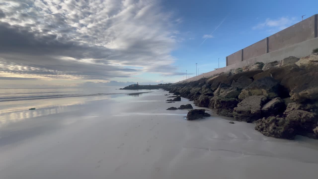 Lighthouse rising above rocks on Barbate beach at sunrise under cloudy sky in Spain, with a concrete wall protecting the coast