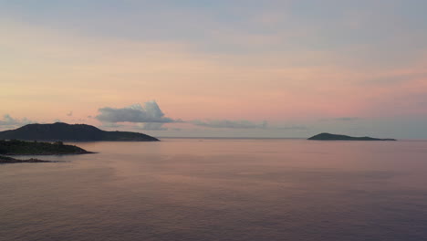 Vista-Infinita-De-Cielos-Rosados-Y-Azules-Durante-La-Puesta-De-Sol-Sobre-Una-Superficie-Lisa-Y-Reflectante-Del-Océano,-Con-Islas-Tropicales-En-La-Distancia