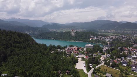 Aerial-flight-over-city-of-Bled-with-forest-and-lake-during-cloudy-day---Silhouette-of-mountain-range-in-background,-Slovenia