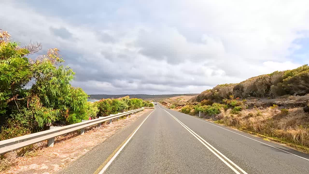 A motorcycle travels along the picturesque Great Ocean Road, showcasing coastal views and lush greenery under a cloudy sky