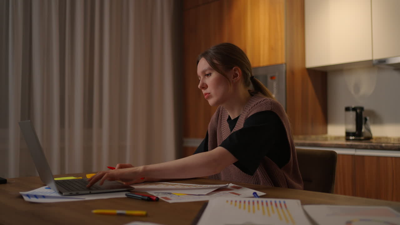 Side view A woman works remotely in a home office at a desk with a laptop and notes data on a graph a business economist analyst. Writes down makes notes. Distance student economist