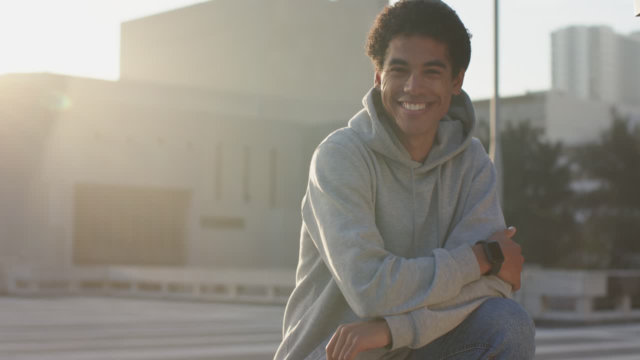 Seated man wearing hoodie, smiling, laughing while sun flare appearing on rooftop ledge, copy space