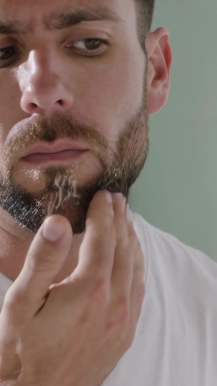 Video verticale: Uomo in maglietta che si tocca il mento con le mani, applica la crema e si strofina la barba al lavandino per la cura.