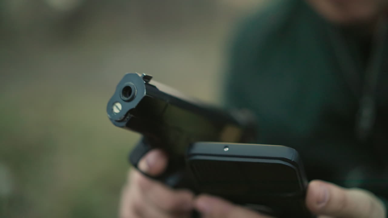 A close-up blur view of a man in a green jacket seated, holding a gun in one hand and a cell phone in the other, as he operates it