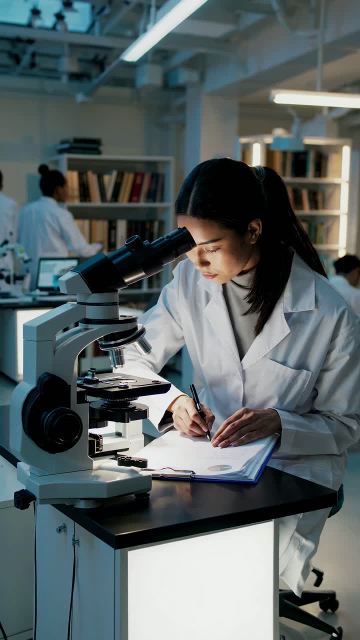 A scientist in a lab coat writes notes beside a microscope. The side angle captures a focused