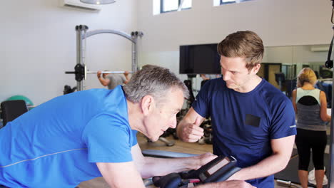 senior man exercising on cycling machine being encouraged by personal trainer in gym