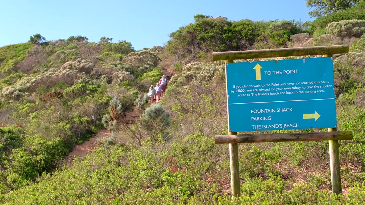 Direction signage with hikers hiking through coastal vegetation on Robberg trail