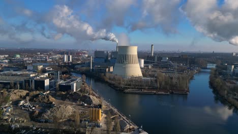 large industrial power plant emitting large amounts of smoke over the city berlin next to a river on a sunny day. smooth aerial view flight ascending drone