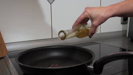 medium handheld shot of a woman's hand with a bottle filled with oil while she adds high quality sunflower oil to a hot pan for cooking in the kitchen for a delicious dinner