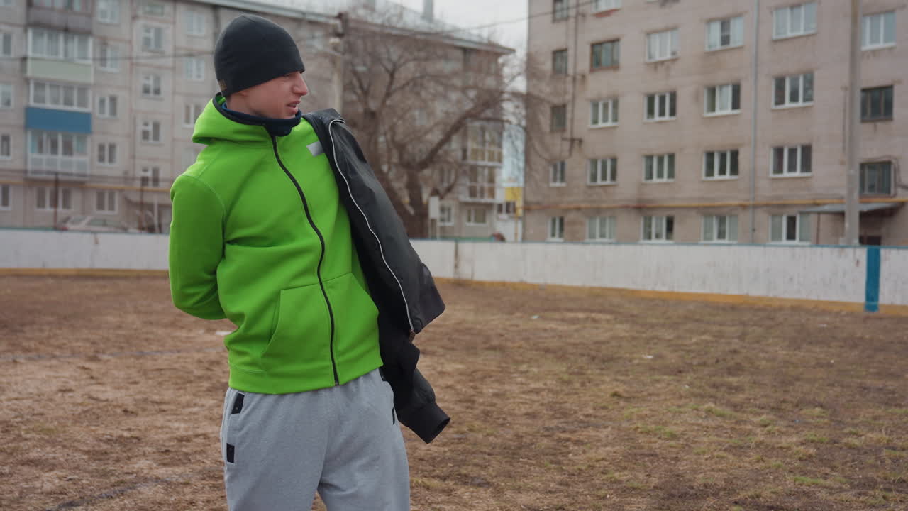Una mujer blanca ofrece una chaqueta a un compañero de equipo blanco en un campo urbano frío, un gesto afectuoso y calor compartido, manos pasando el abrigo, apoyo amistoso después del entrenamiento, espíritu comunitario en un partido vecinal
