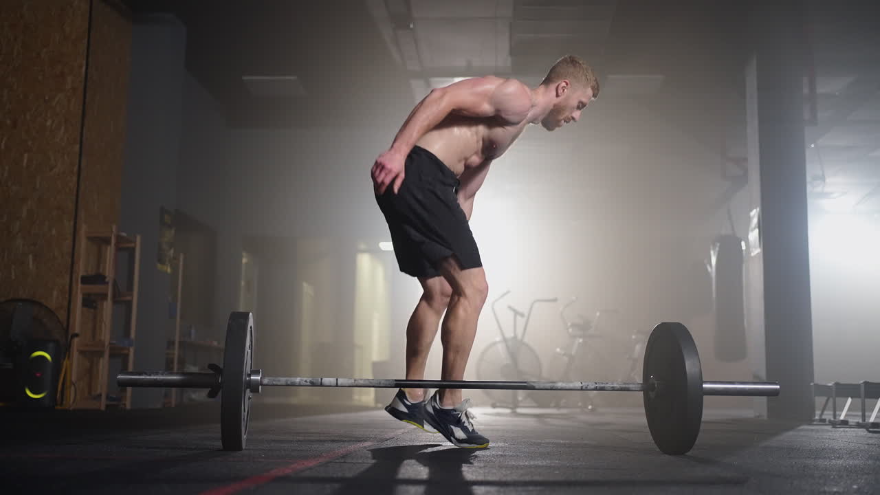 Un giovane senza maglietta in slow motion salta un burpee sopra un bilanciere in palestra in controluce