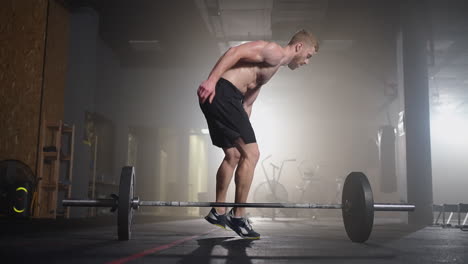 a young man without a shirt in slow motion jumps a burpee over a barbell in the gym in backlight