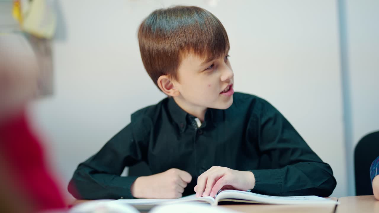 Cute boy in the classroom. Kid is sitting at the desk and listening to the teacher at primary school. Clever boy in black shirt talking during the lesson.