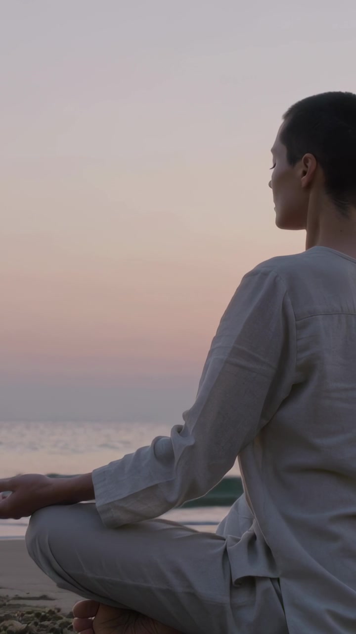 Video verticale: Donna che medita all'alba in posizione yoga sulla spiaggia, che guarda il mare e il cielo tenue, con spazio per il testo.