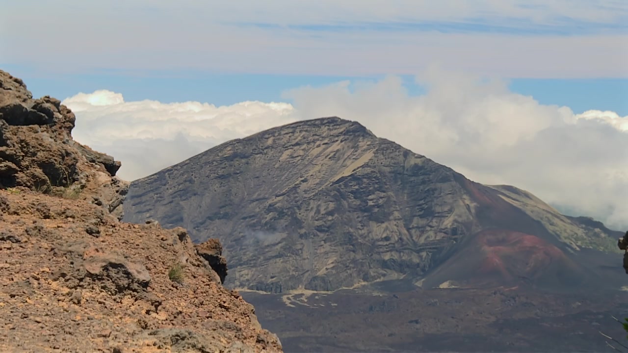 Ampia ripresa diurna della scoscesa cima vulcanica di Haleakala, drammatiche formazioni rocciose e paesaggio craterico sotto un cielo luminoso con nuvole alla deriva. Nessuna persona visibile.