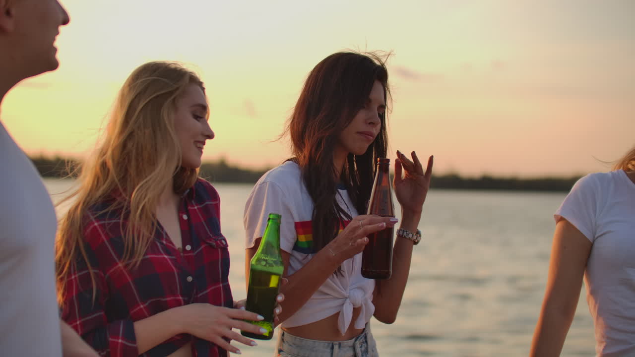 tres chicas jóvenes con cabello largo están bailando en la fiesta al aire libre en la playa con cerveza. están moviendo sus cuerpos jóvenes en la costa del lago y teniendo un gran momento al atardecer con amigos.