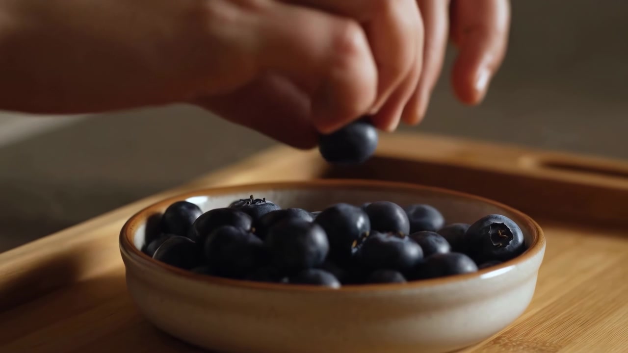 Un primo piano mostra mani umane che sollevano con cura un mirtillo da una piccola ciotola di ceramica piena di bacche scure su un tavolo di legno, con luce calda e soffusa, sfondo attenuato, texture lucida e un'atmosfera intima.