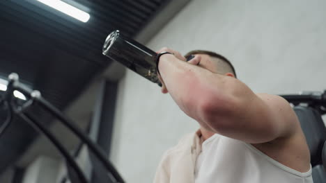 young man seated on weight bench in modern gym taking refreshing water drink after intense chest press exercise pushing heavy loads resting between sets capturing recovery focus moment
