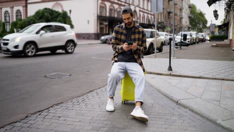 a man sitting on his yellow suitcase on a city street, looking at his phone