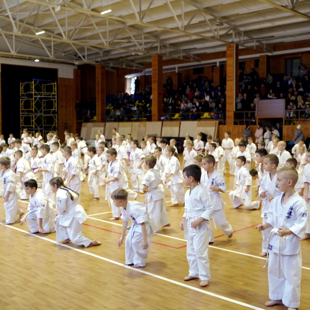 Children studying karate stand in rows in a big gym. Kids sit down on the floor together. Audience sitting at background
