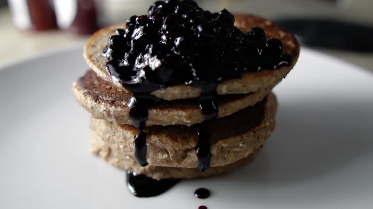 Blueberry sauce dripping from four stacked oat pancakes on a white plate, SLOW MOTION