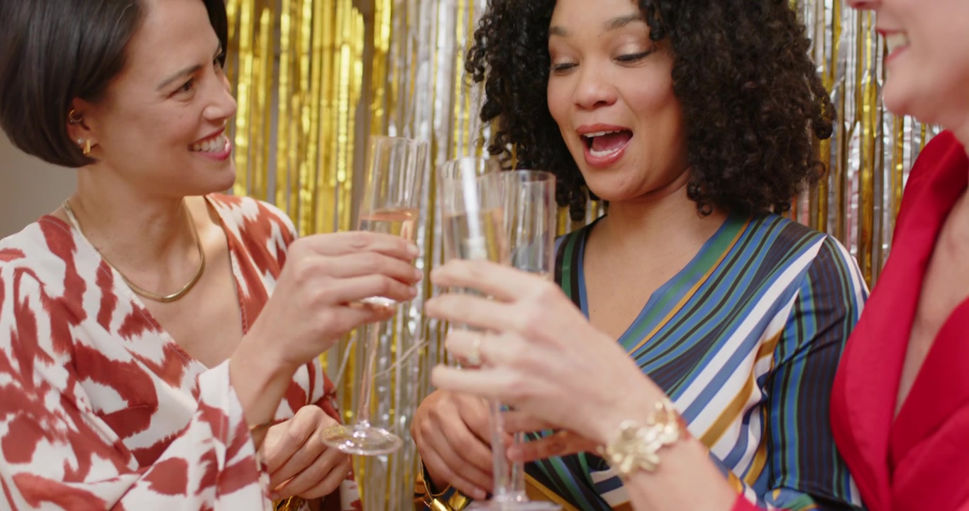 Diverse female friends celebrating, raising champagne flutes and blowing gold noisemakers at party